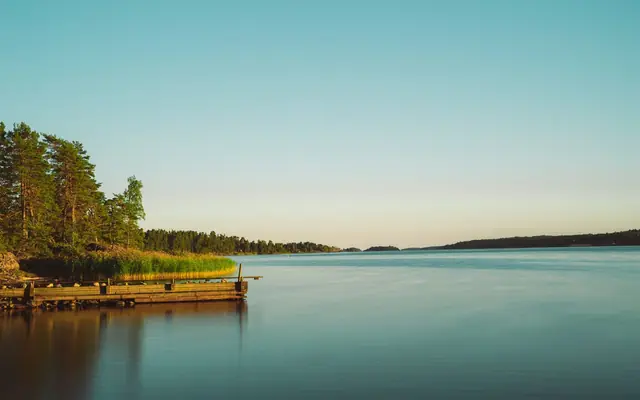 Serene view of a calm lake with lush green forests and wooden dock under a clear sky at sunset.