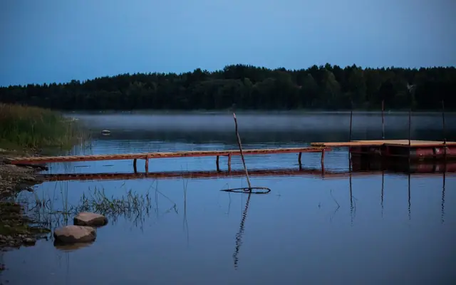 Tranquil lakeside scene with a wooden dock reflecting in the calm water at dusk under a clear sky.