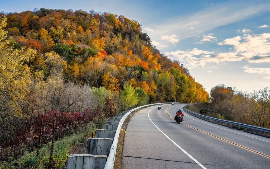 Motorcyclists ride along a scenic road surrounded by vibrant autumn foliage.