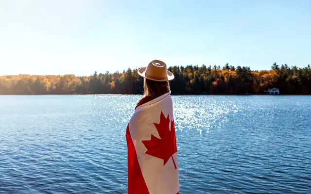 A woman wrapped in a Canadian flag stands by a scenic Ontario lake during fall.