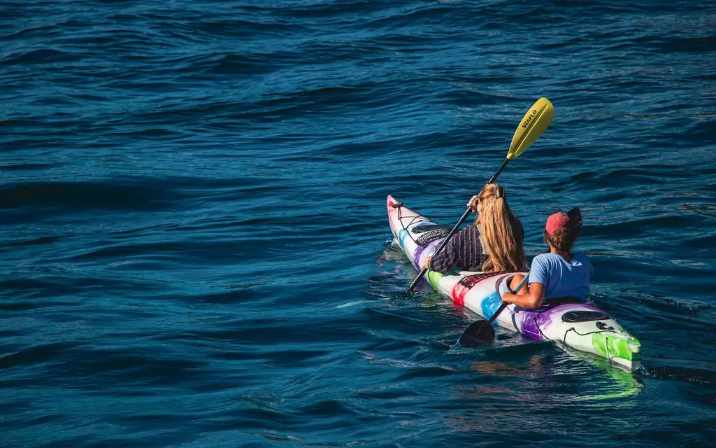 Two women paddle a colorful kayak on a calm lake during a sunny summer day, enjoying outdoor recreation.