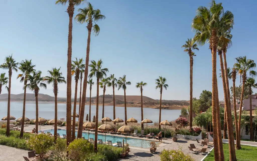 Tranquil resort setting featuring palm trees, a pool, and a stunning lake view under a clear blue sky.